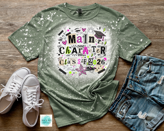 Green t-shirt with 'Main Character Class of 2026' design on a wooden surface with white sneakers and denim shorts.
