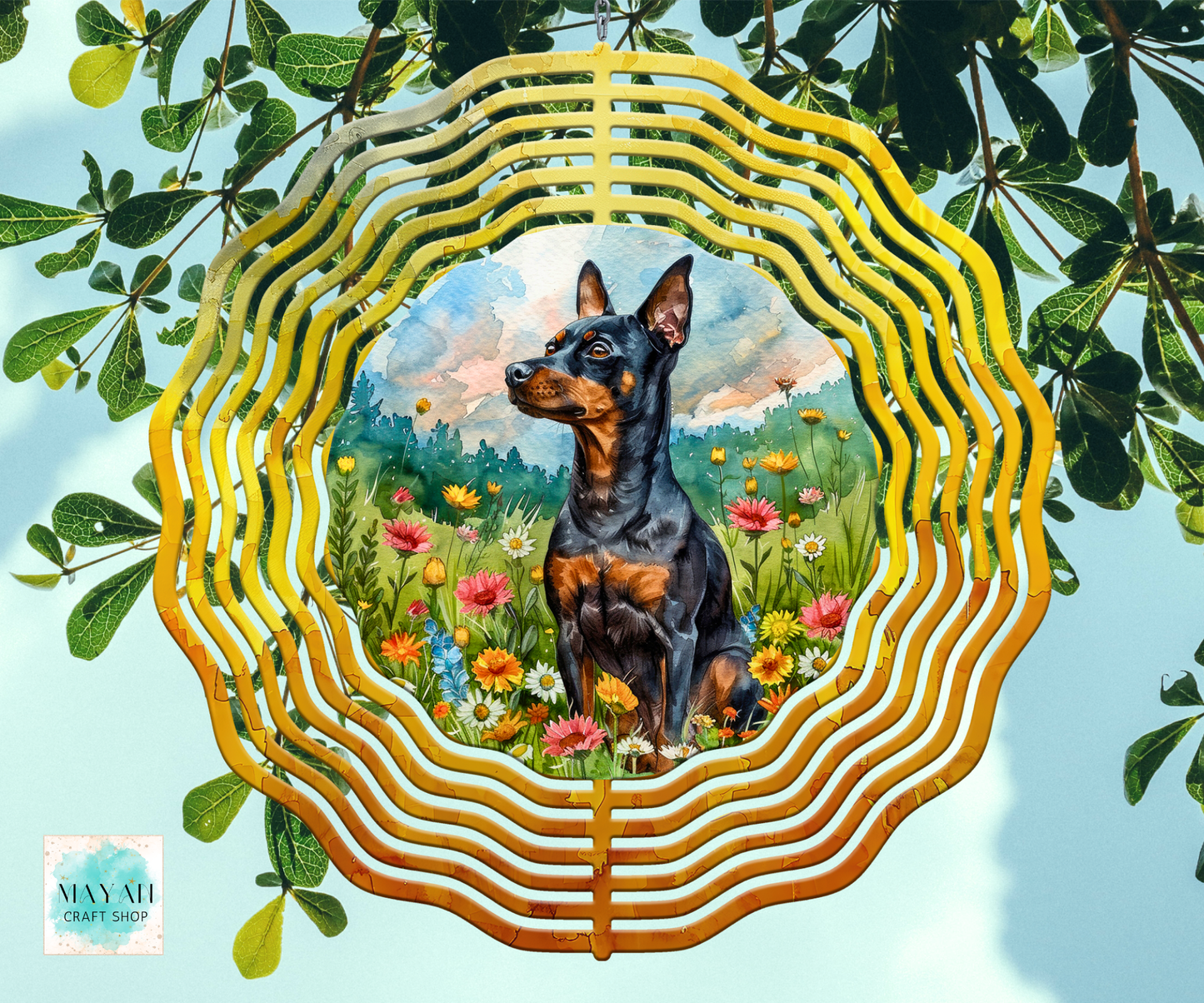 A wind spinner, featuring a painted scene of a black and tan dog sitting in a flower field. The piece is photographed hanging against a light blue sky with green leaves around it.