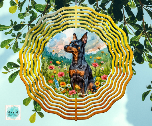 A wind spinner, featuring a painted scene of a black and tan dog sitting in a flower field. The piece is photographed hanging against a light blue sky with green leaves around it.