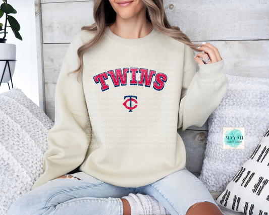 Person wearing a sand-colored sweatshirt with baseball themed text and logo, sitting on a couch.
