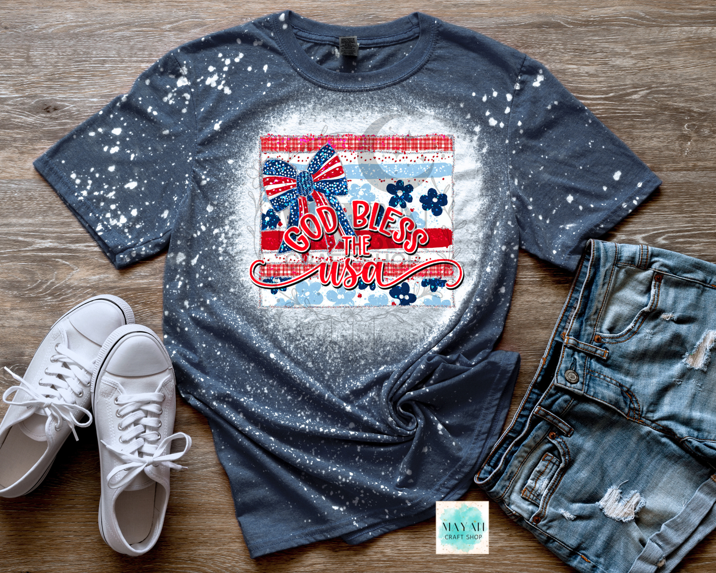 Blue bleached t-shirt with 'God Bless the USA' graphic on a wooden surface with white sneakers and denim shorts.