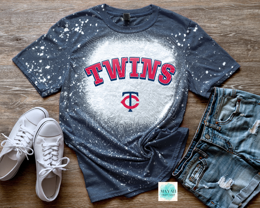 Blue baseball t-shirt with white splatter design, white sneakers, and denim shorts on a wooden surface.