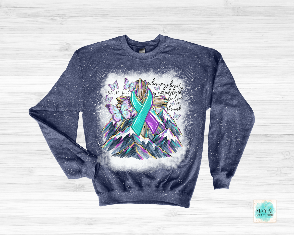 A navy blue crewneck sweatshirt with a distressed texture, featuring a colorful graphic of a teal and purple ribbon over mountains, butterflies, and a text on a light wood background.