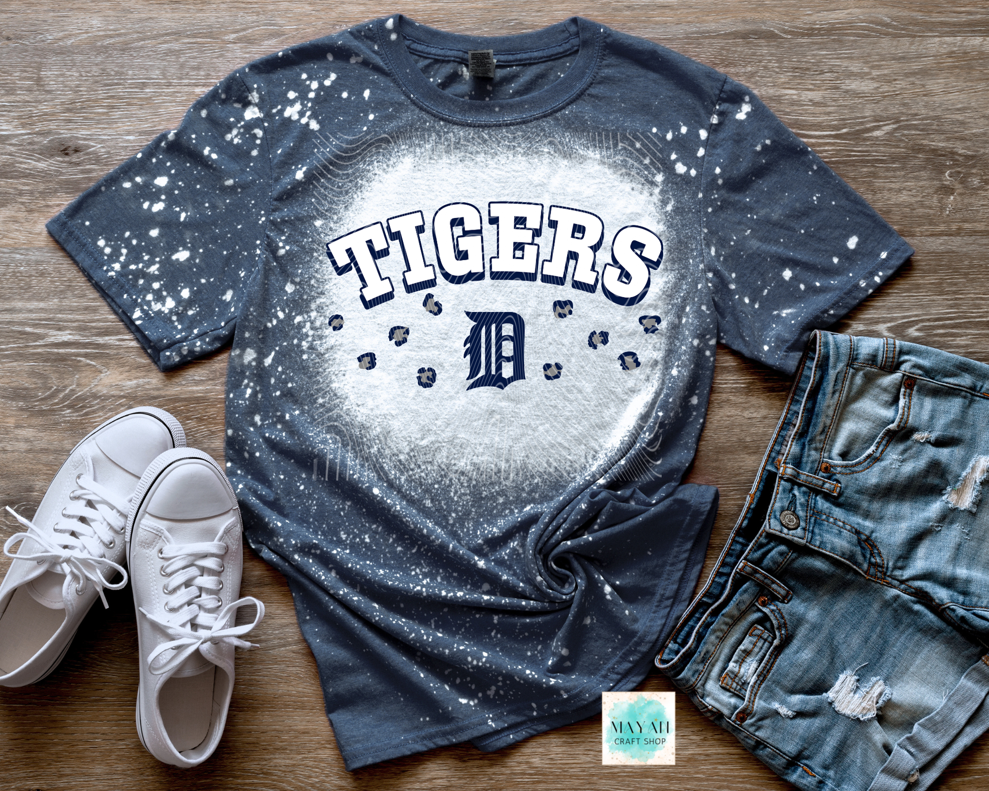 Blue baseball t-shirt with white splatter design, white sneakers, and denim shorts on a wooden surface.