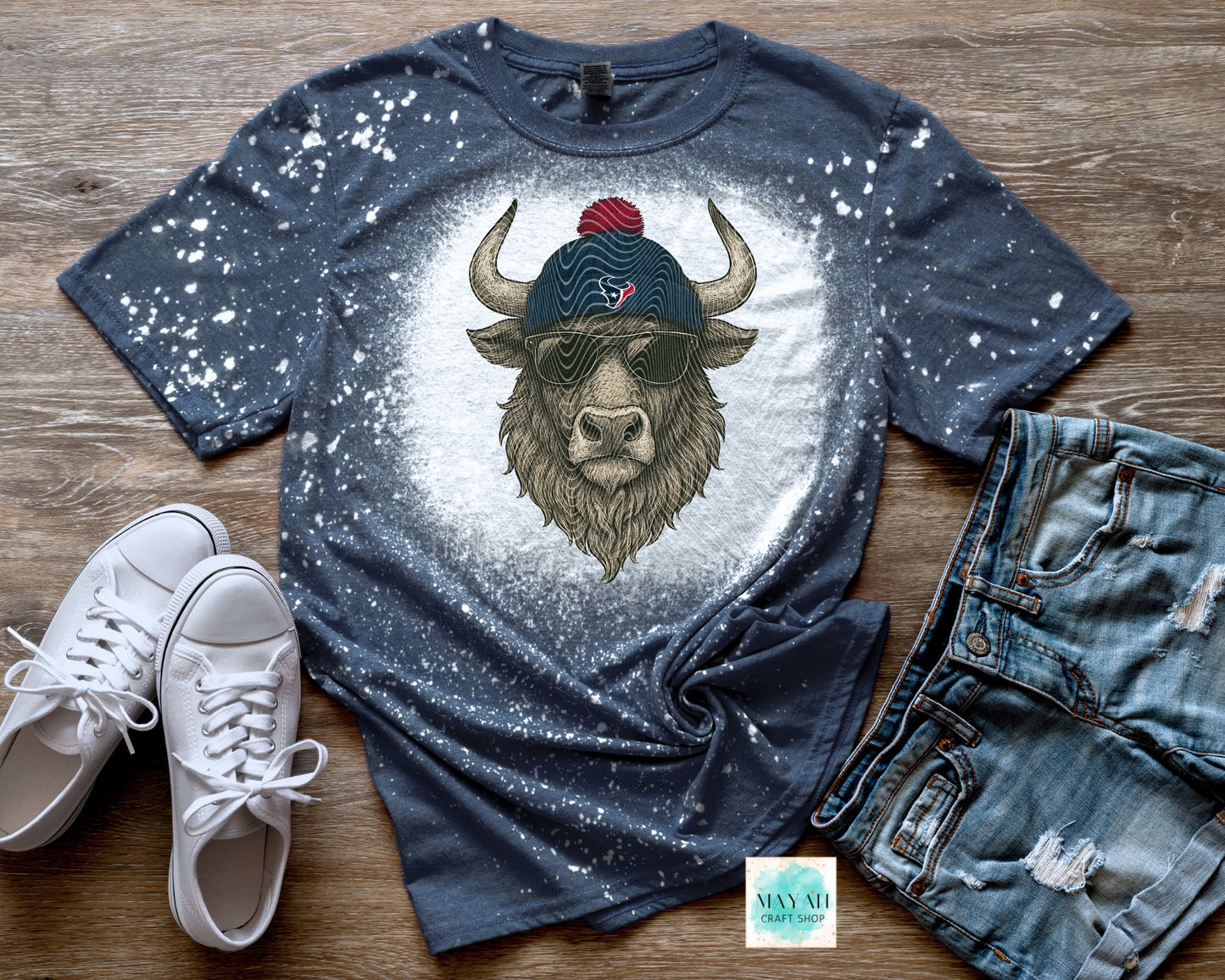 Blue t-shirt with a bull graphic wearing sunglasses and a beanie, paired with white sneakers and jeans on a wooden surface.