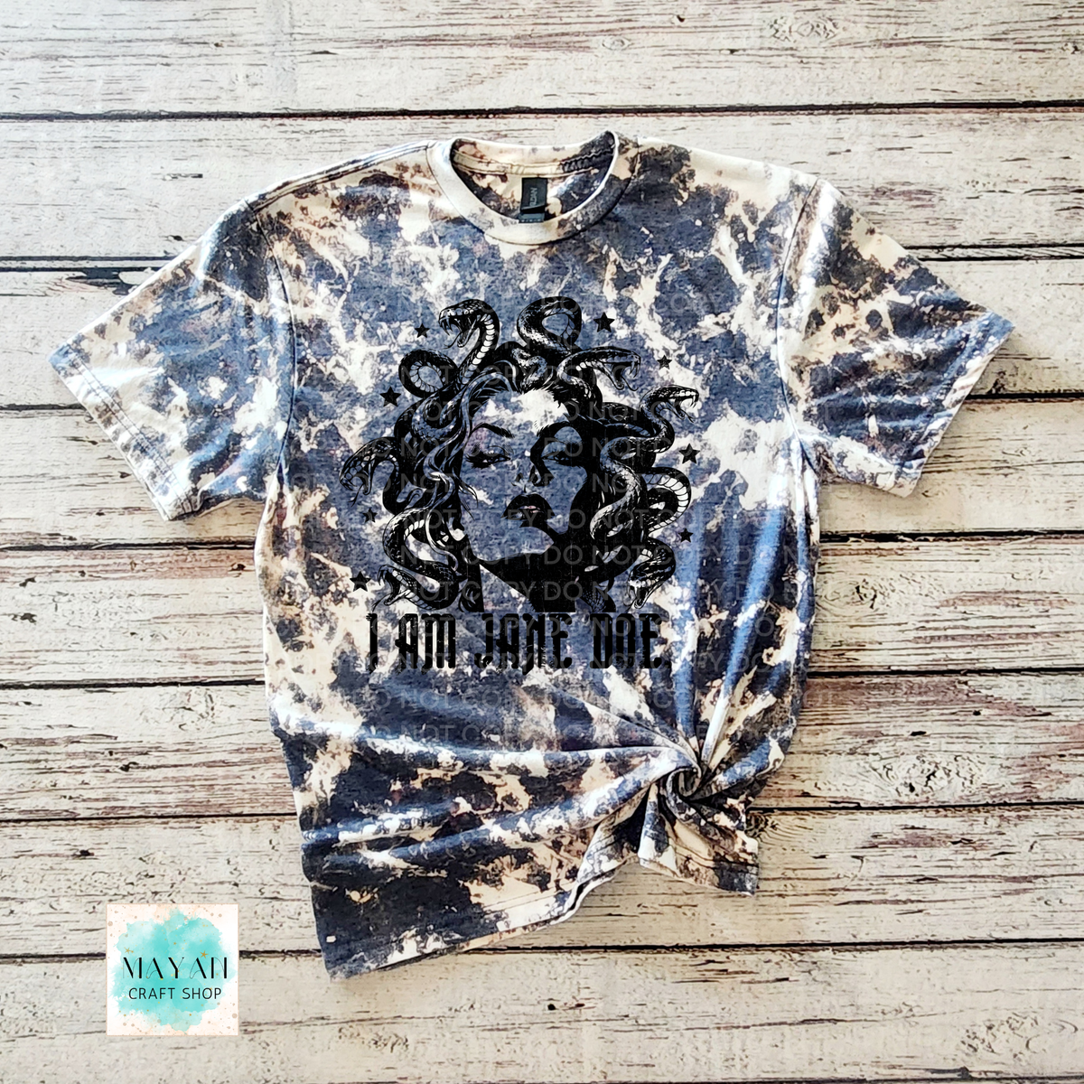 A short-sleeve crewneck heather navy bleached t-shirt, featuring a black graphic of Medusa and the text 'I AM JANE DOE' on the front. The shirt is styled flat on a rustic wooden plank background with a knot at the bottom hem.