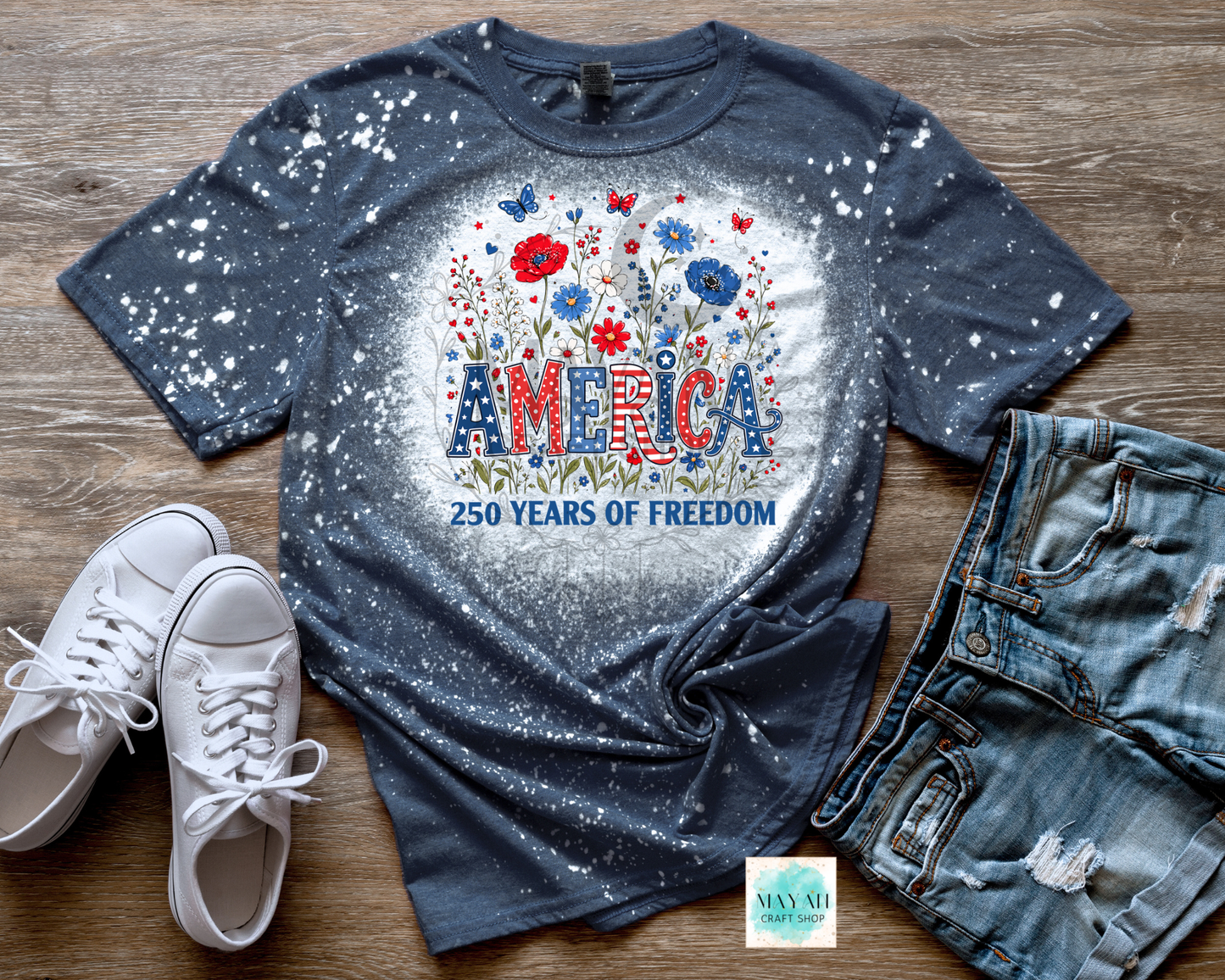 Blue graphic t-shirt with 'America 250 Years of Freedom' design on a wooden surface with white sneakers and denim shorts.