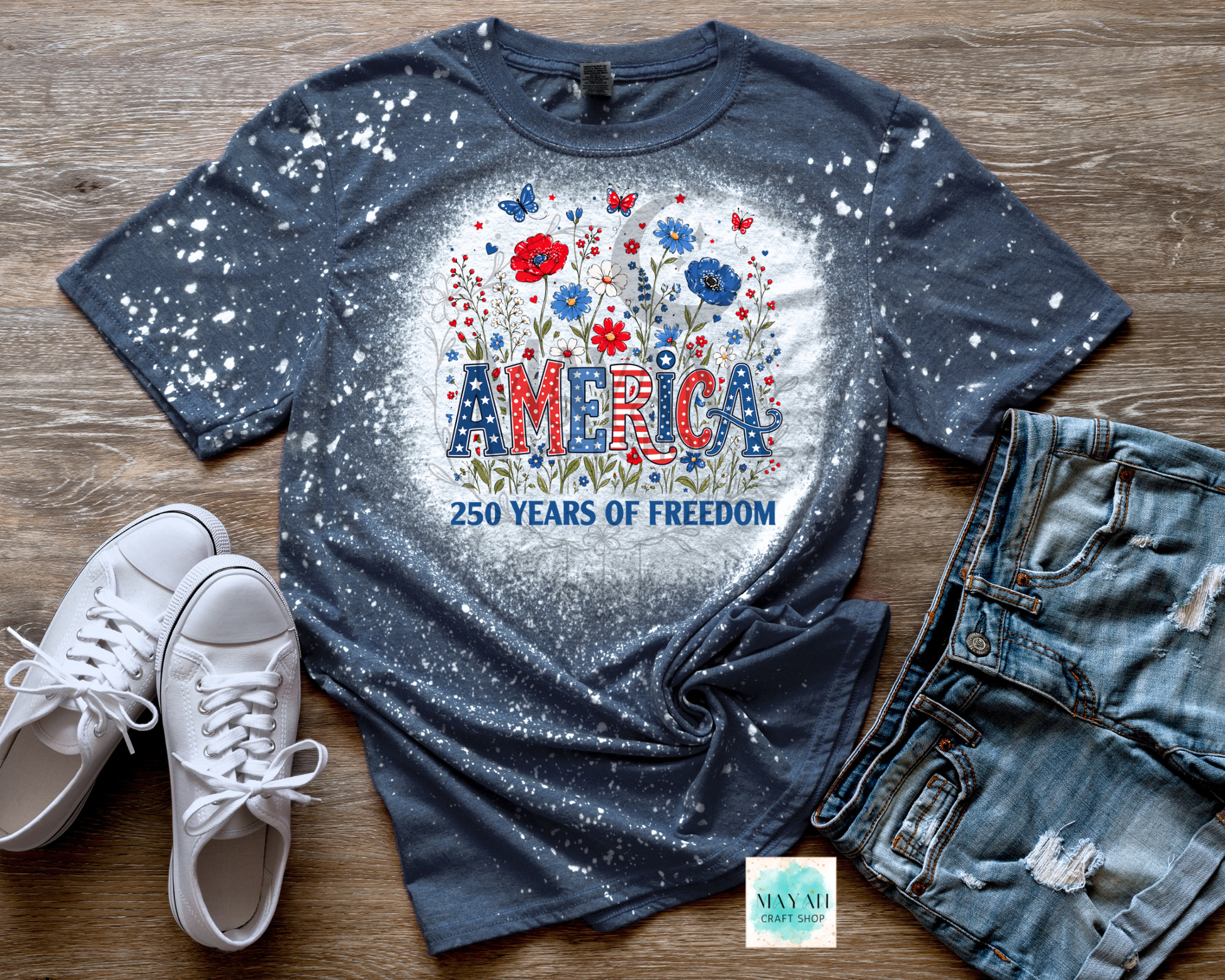 Blue graphic t-shirt with 'America 250 Years of Freedom' design on a wooden surface with white sneakers and denim shorts.