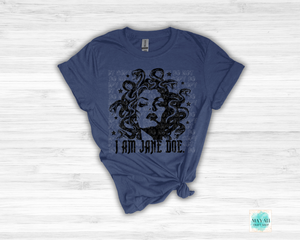 A navy blue graphic t-shirt featuring a black distressed print of Medusa with snakes and the text 'I AM JANE DOE', styled flat on a white wood background.