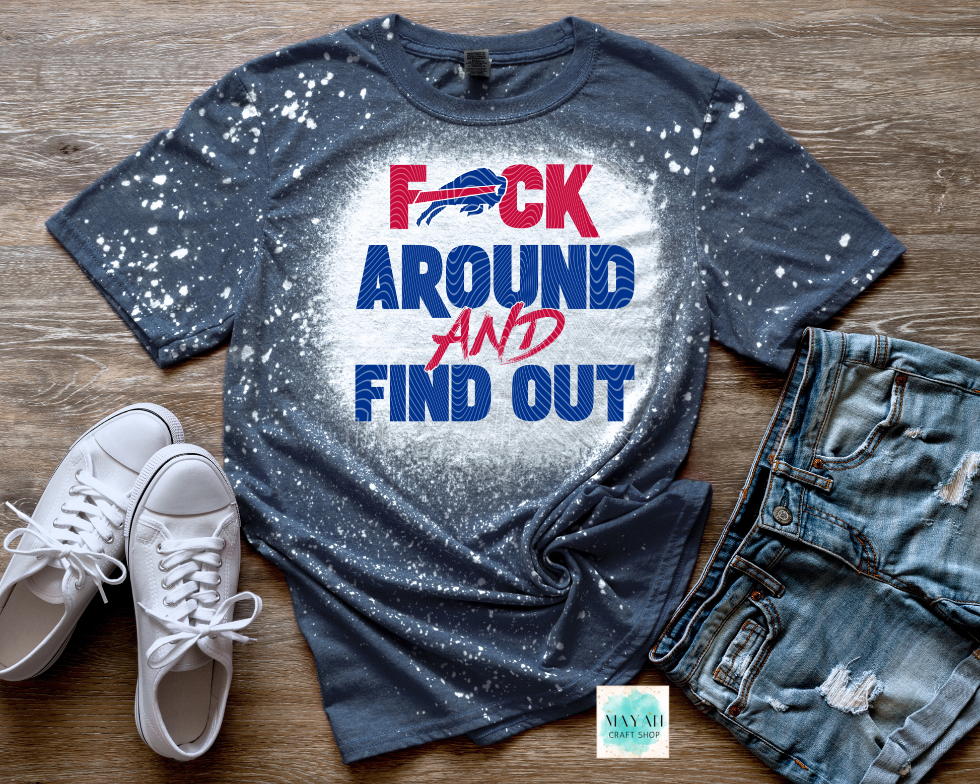 Blue t-shirt with graphic design and text on a wooden surface with shoes and jeans.