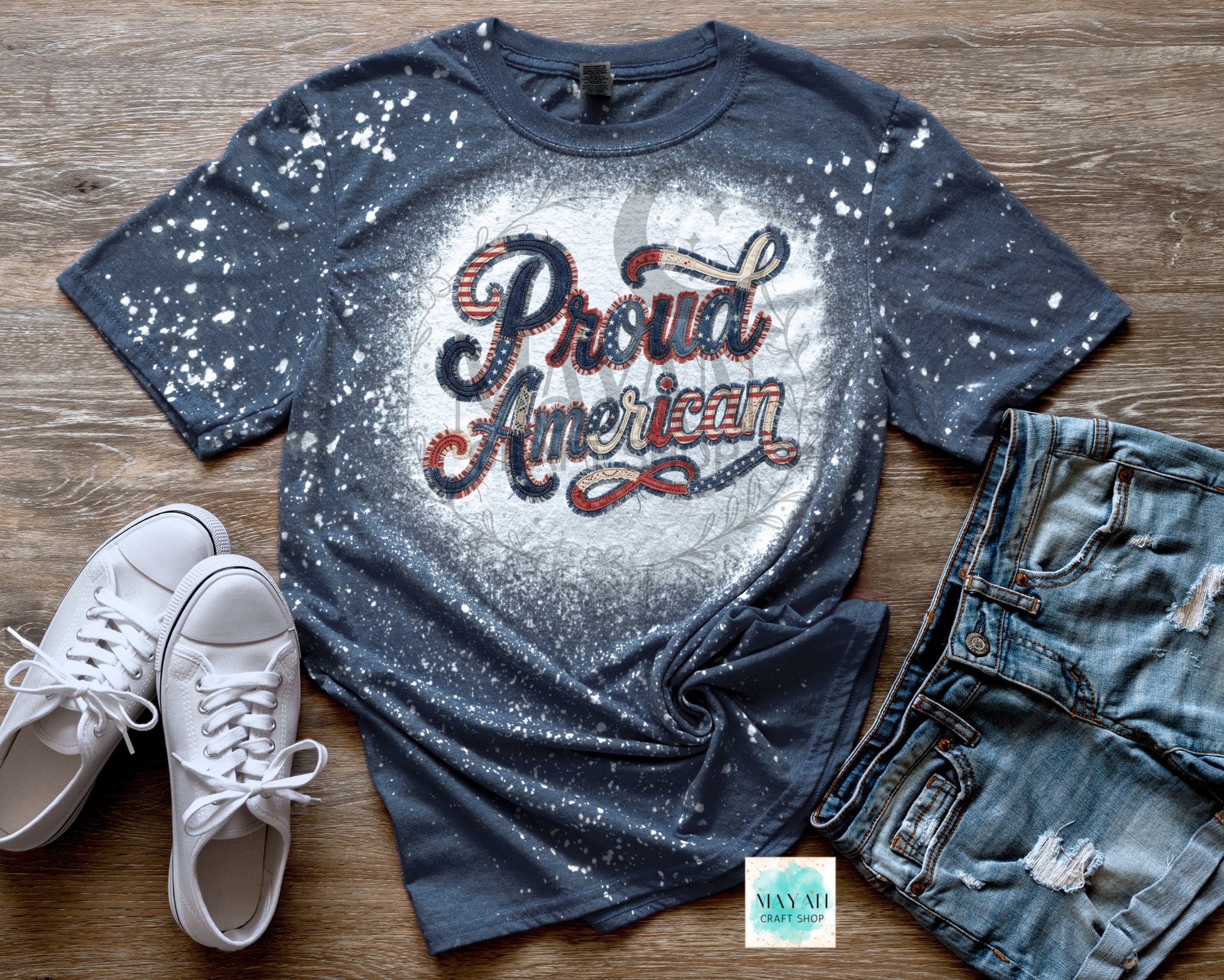 Blue splatter paint t-shirt with 'Proud American' text, white sneakers, and denim shorts on a wooden surface.