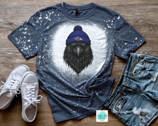 Blue t-shirt with a graphic of a bird wearing a blue beanie, paired with white sneakers and jeans on a wooden surface.