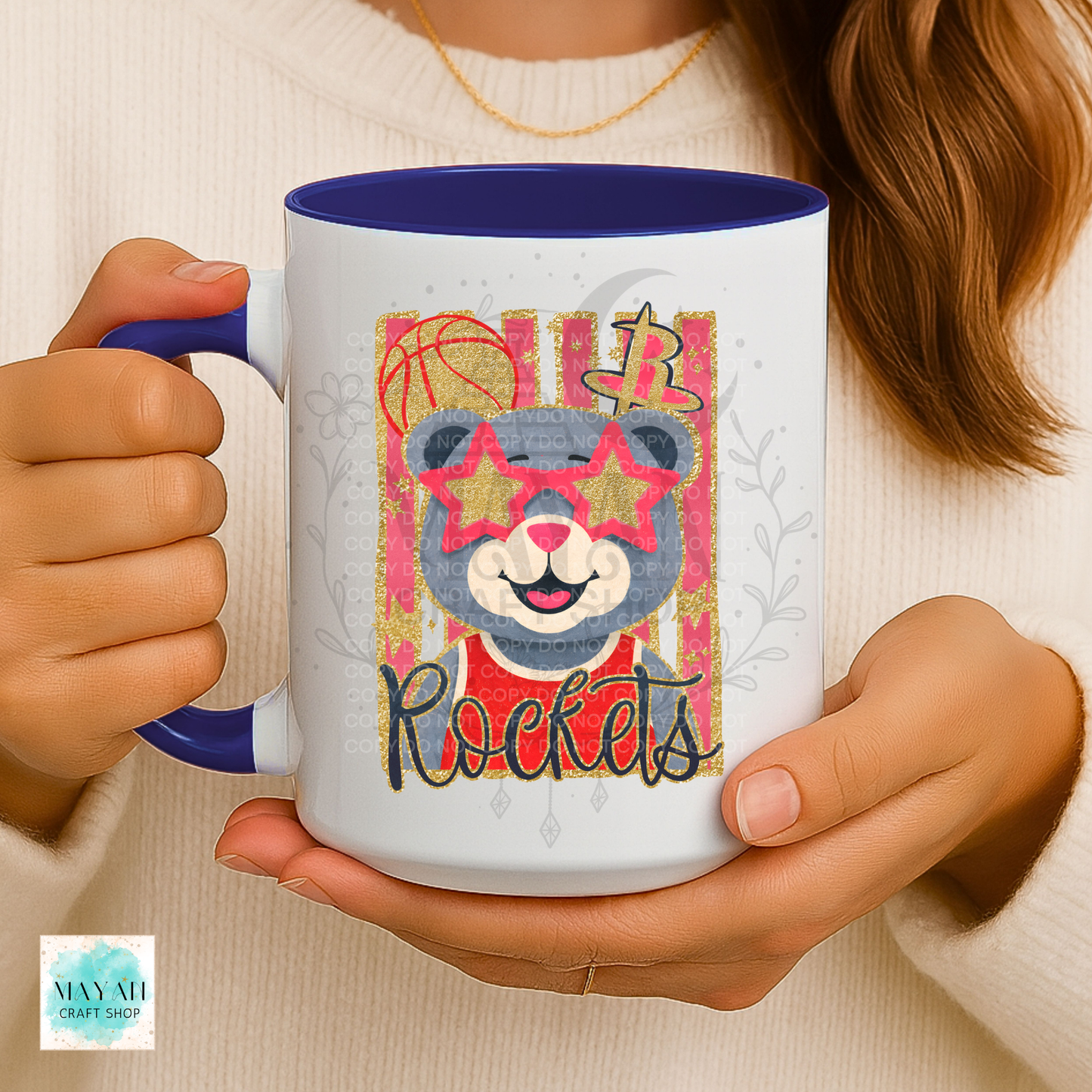 A person holds a white ceramic mug with a blue interior and handle. The mug features a colorful graphic of a bear wearing star-shaped sunglasses with a word below it.