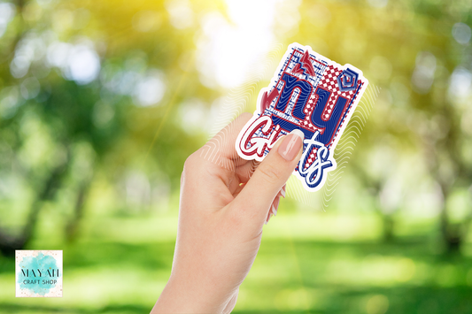 A hand holds a die-cut sticker that reads New York in red, white, and blue with a plaid pattern, set against a blurred green outdoor background.