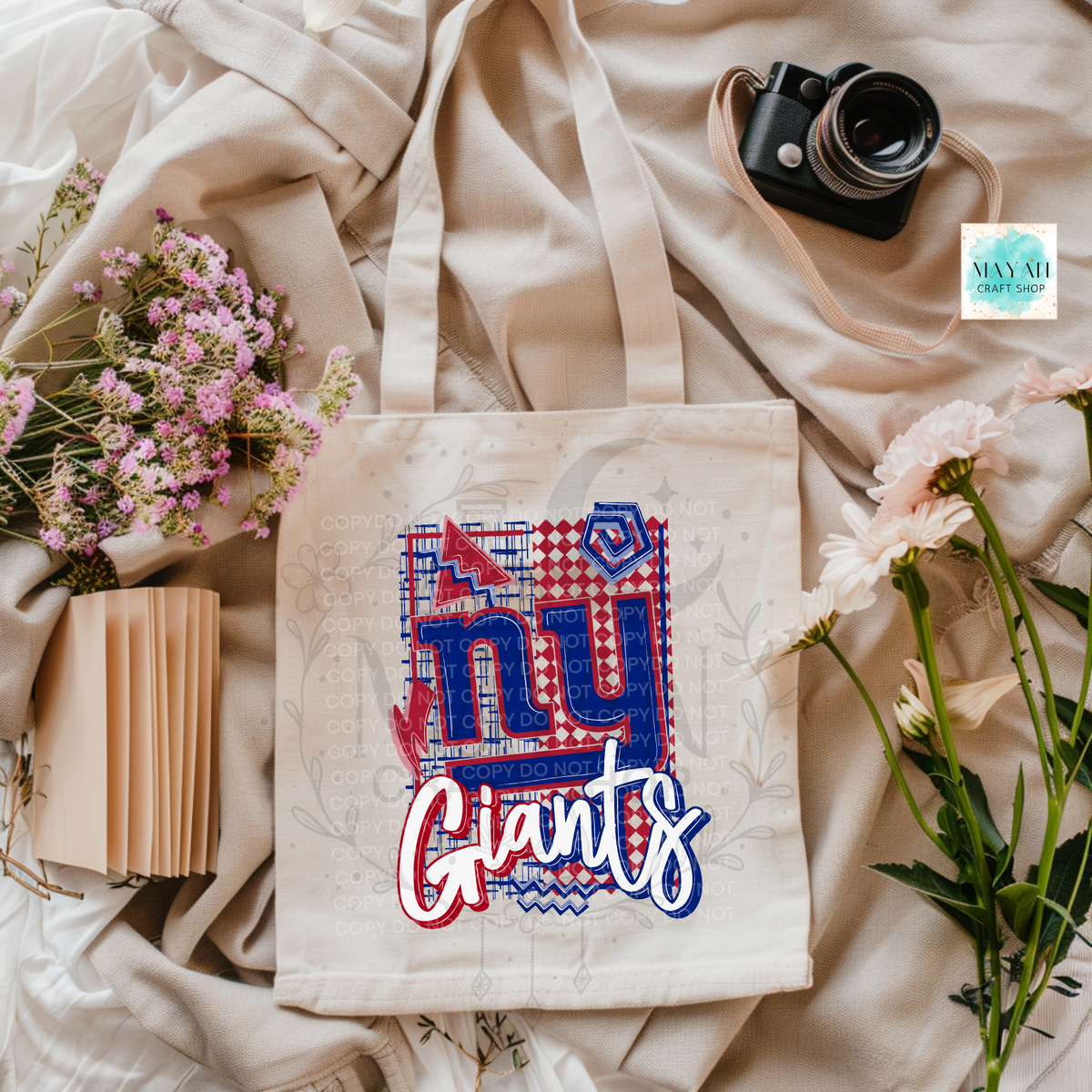 A cream canvas tote bag featuring a red, white, and blue football logo design, styled in a flat-lay with flowers, a camera, and a notebook on a beige fabric background.