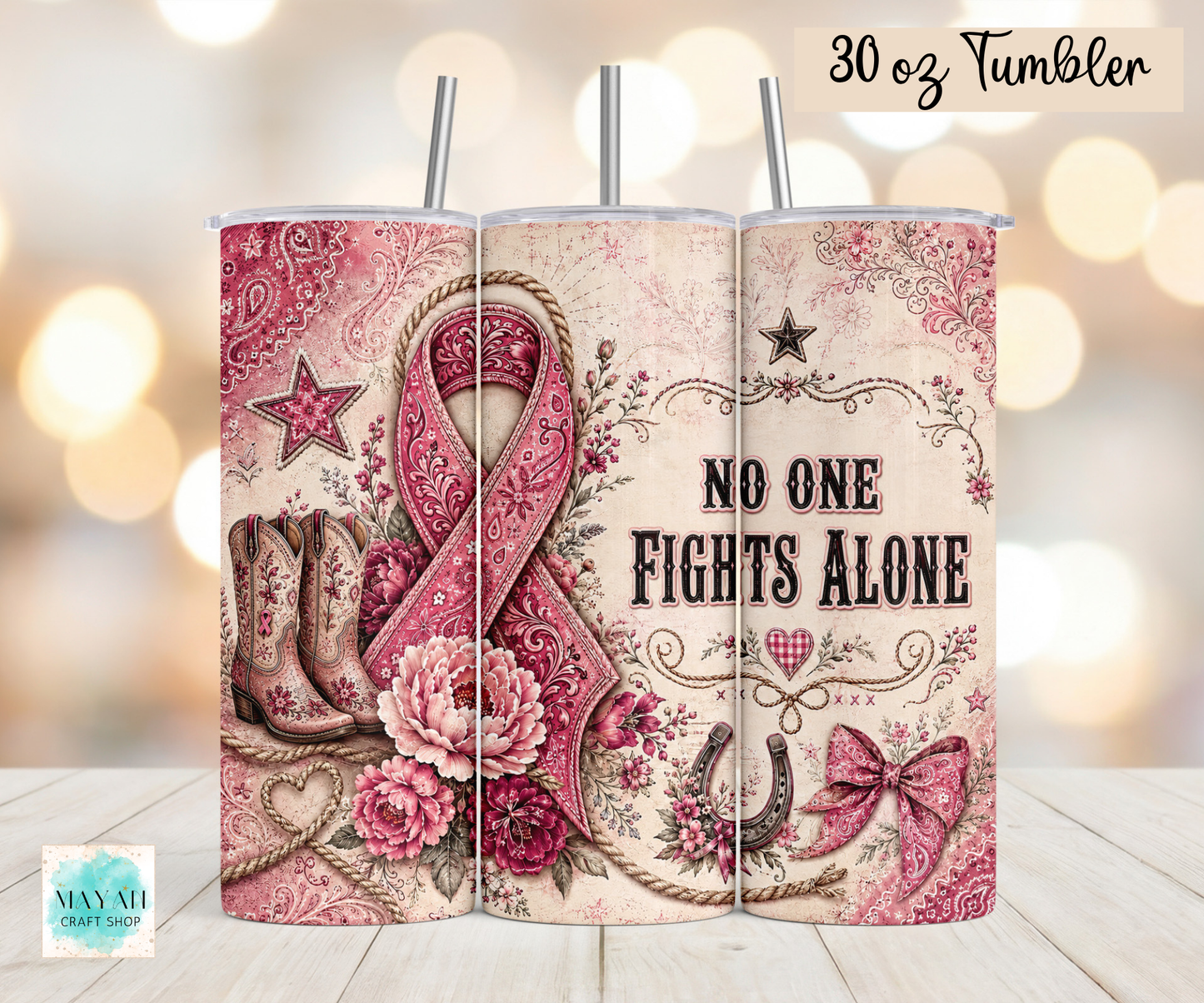 A 30 oz stainless steel tumbler shown from the back, featuring a western-themed design with a pink awareness ribbon, cowboy boots, flowers, bows, and the text 'NO ONE FIGHTS ALONE'. The tumbler has two metal straws and is displayed on a light wood surface with a warm bokeh background.