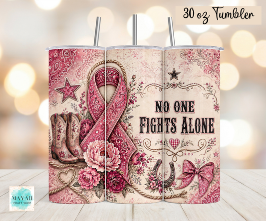 A 30 oz stainless steel tumbler shown from the back, featuring a western-themed design with a pink awareness ribbon, cowboy boots, flowers, bows, and the text 'NO ONE FIGHTS ALONE'. The tumbler has two metal straws and is displayed on a light wood surface with a warm bokeh background.