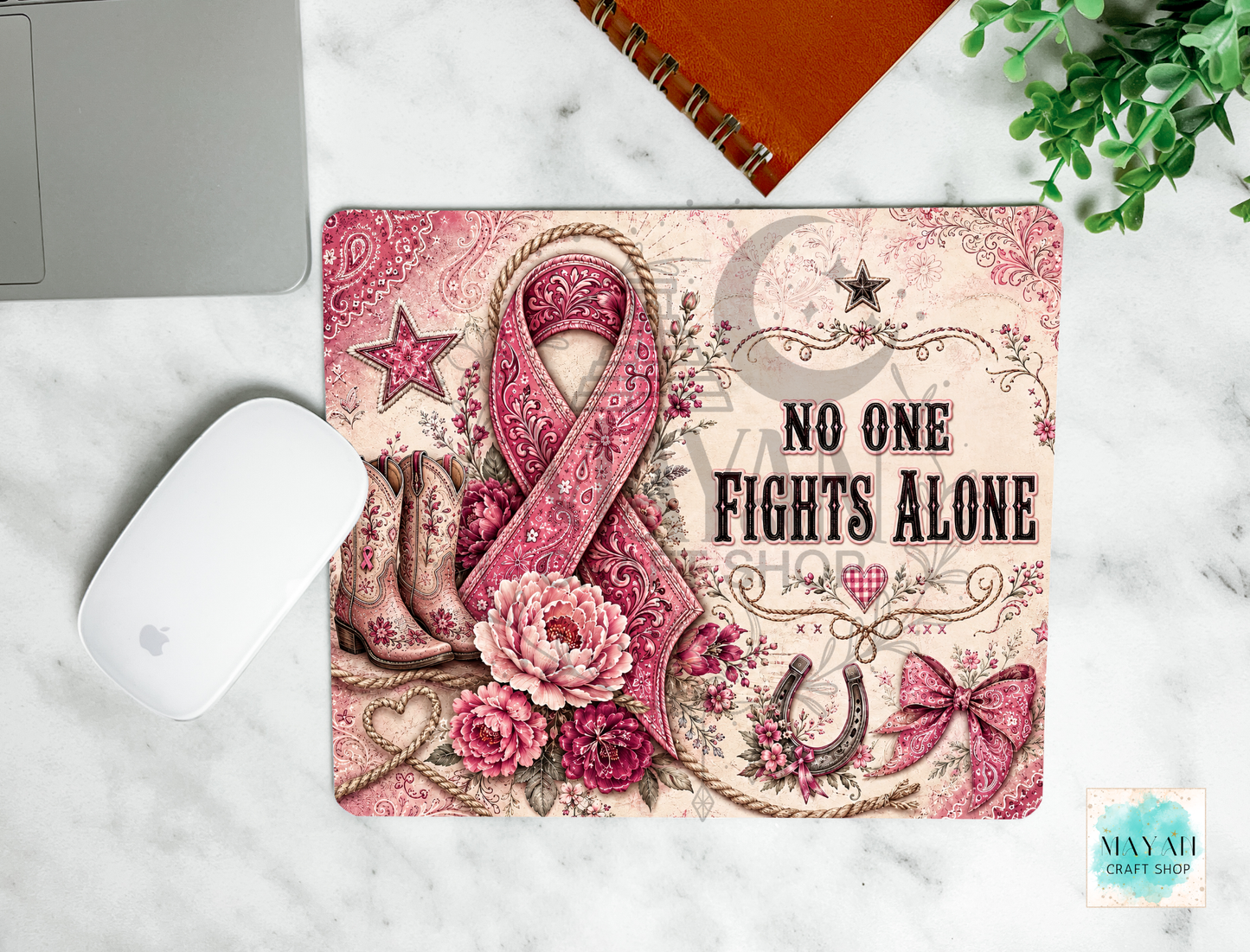 A mouse pad with a western-style pink and beige design featuring a ribbon, flowers, stars, and the text 'NO ONE FIGHTS ALONE,' shown on a white marble surface with a mouse, laptop edge, notebook, and plant nearby.
