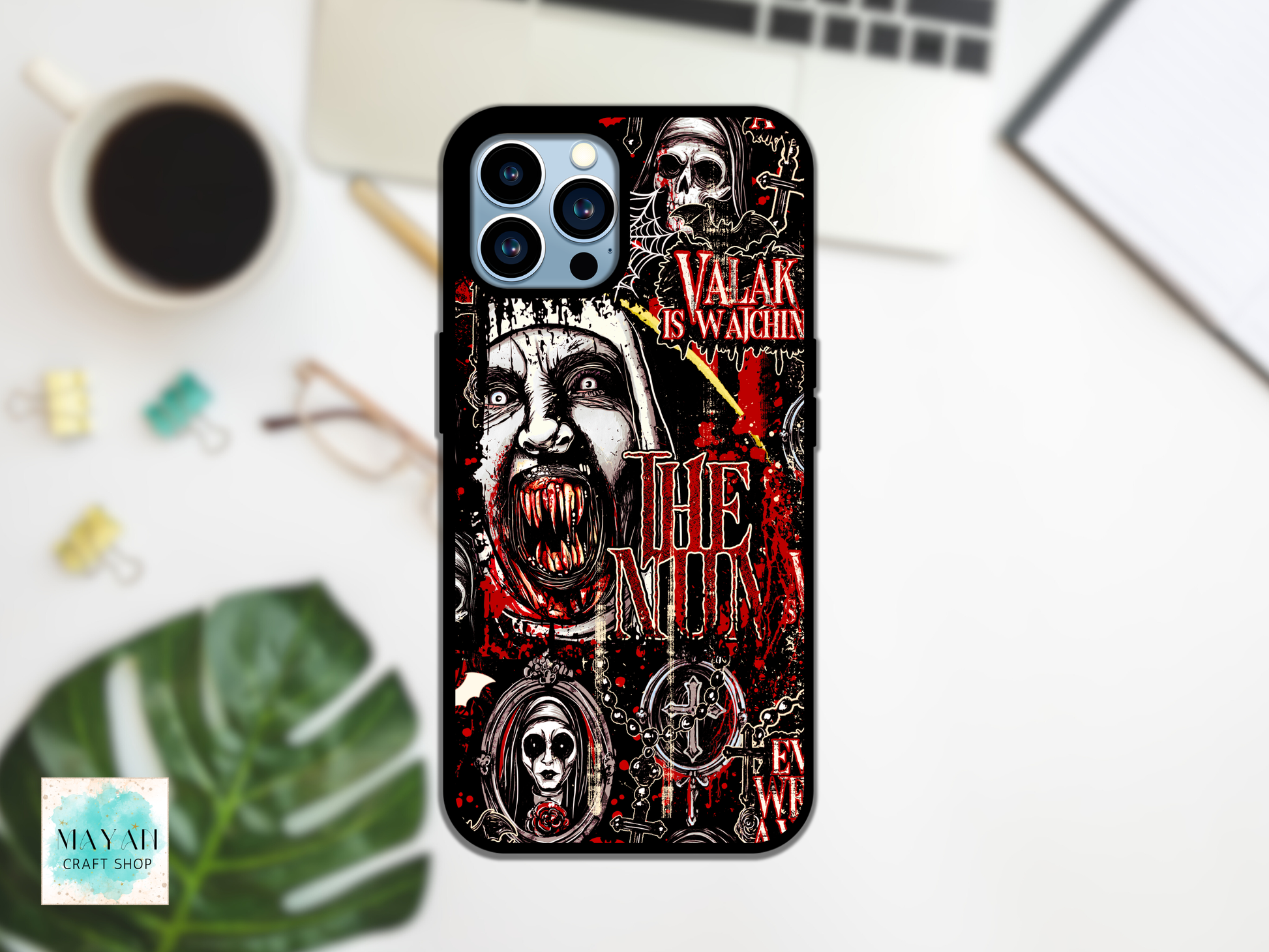A black phone case with a gothic horror graphic print featuring a screaming face, skulls, and red text is displayed on a white desk flat-lay with a coffee cup, laptop, and green leaf.