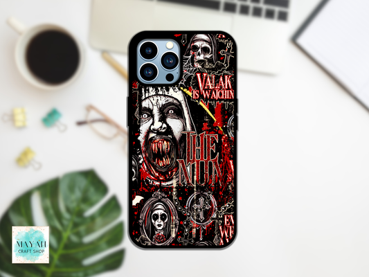 A black phone case with a gothic horror graphic print featuring a screaming face, skulls, and red text is displayed on a white desk flat-lay with a coffee cup, laptop, and green leaf.