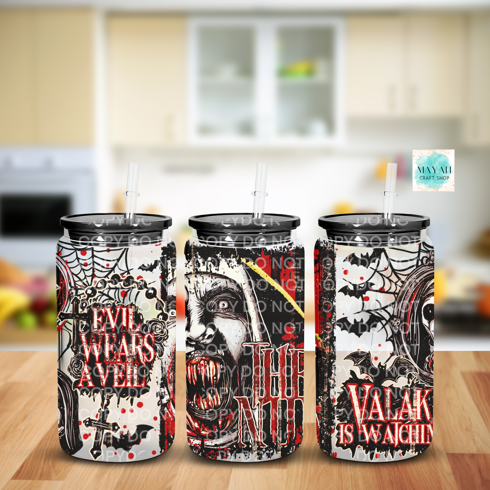Three Halloween-themed frosted glass tumblers with black lids and clear straws, displayed side-by-side on a wooden table with a blurred kitchen background. The tumblers feature spooky graphics including a skull, bats, and spiderwebs.