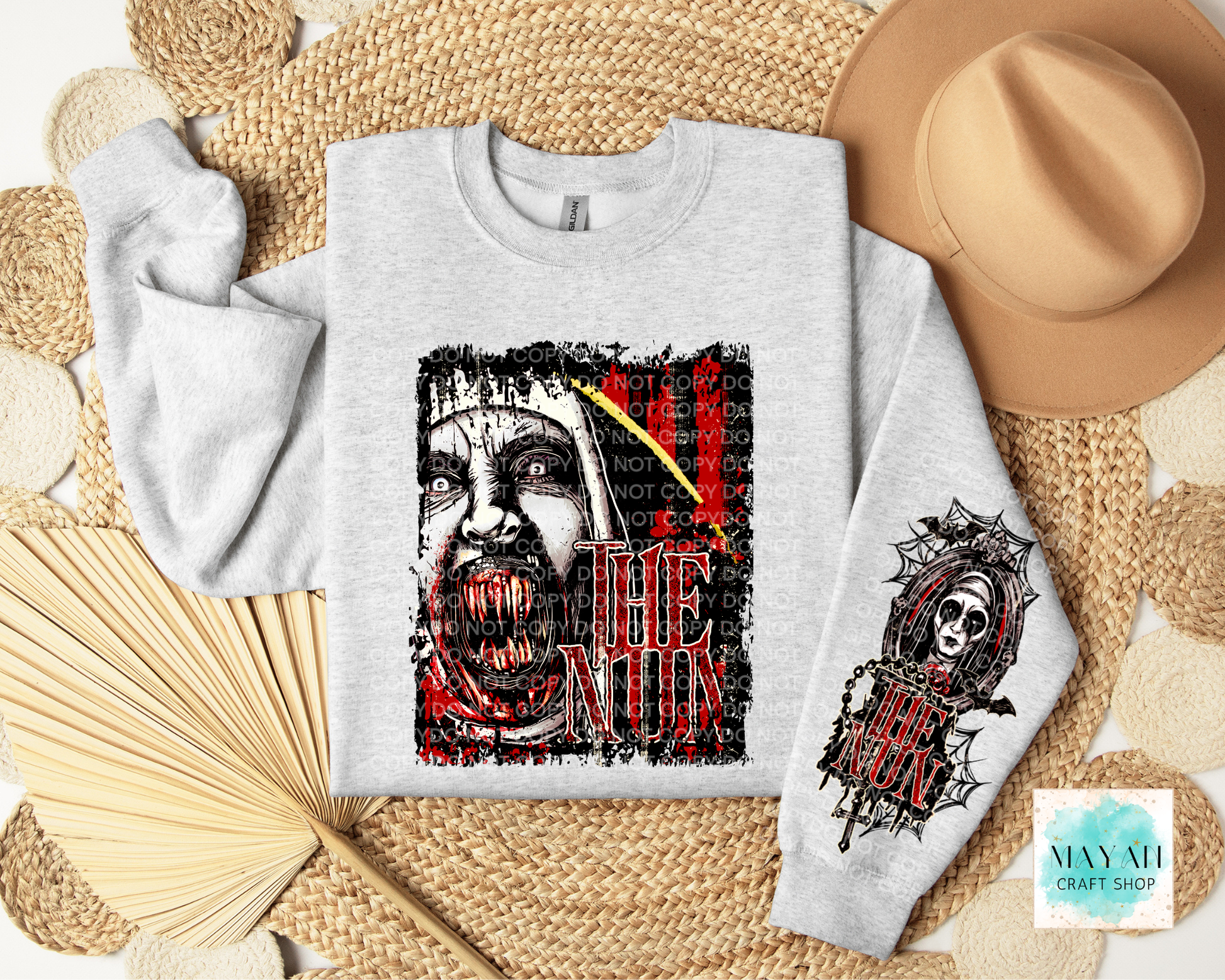A light gray crewneck sweatshirt with a distressed horror graphic of a screaming face in red and black, styled in a flat lay with a tan hat, woven mat.