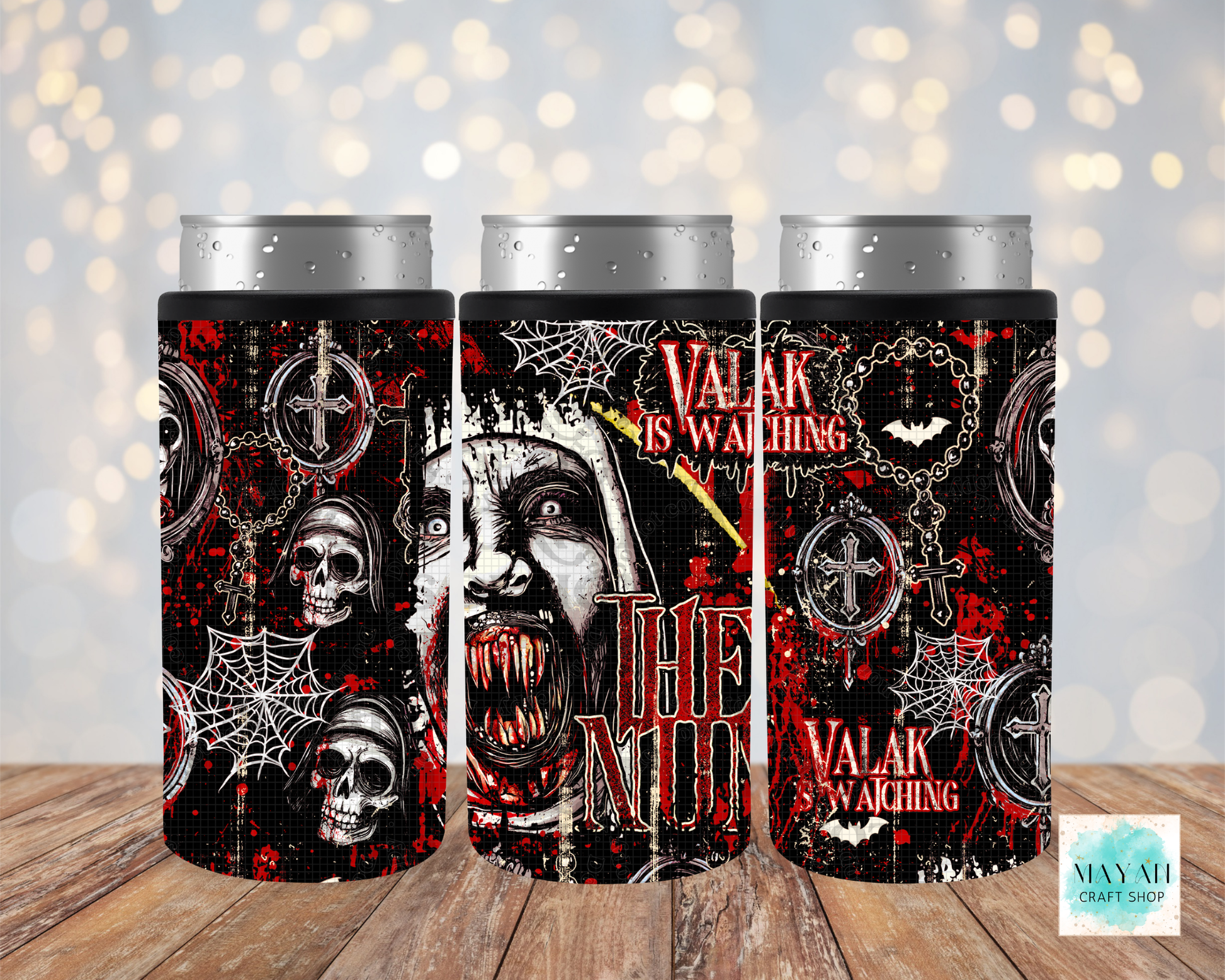 Three slim can coolers with silver aluminum can tops, featuring a gothic horror design with skulls, caskets, cobwebs, on a black background, displayed on a wooden surface with a soft bokeh light background.