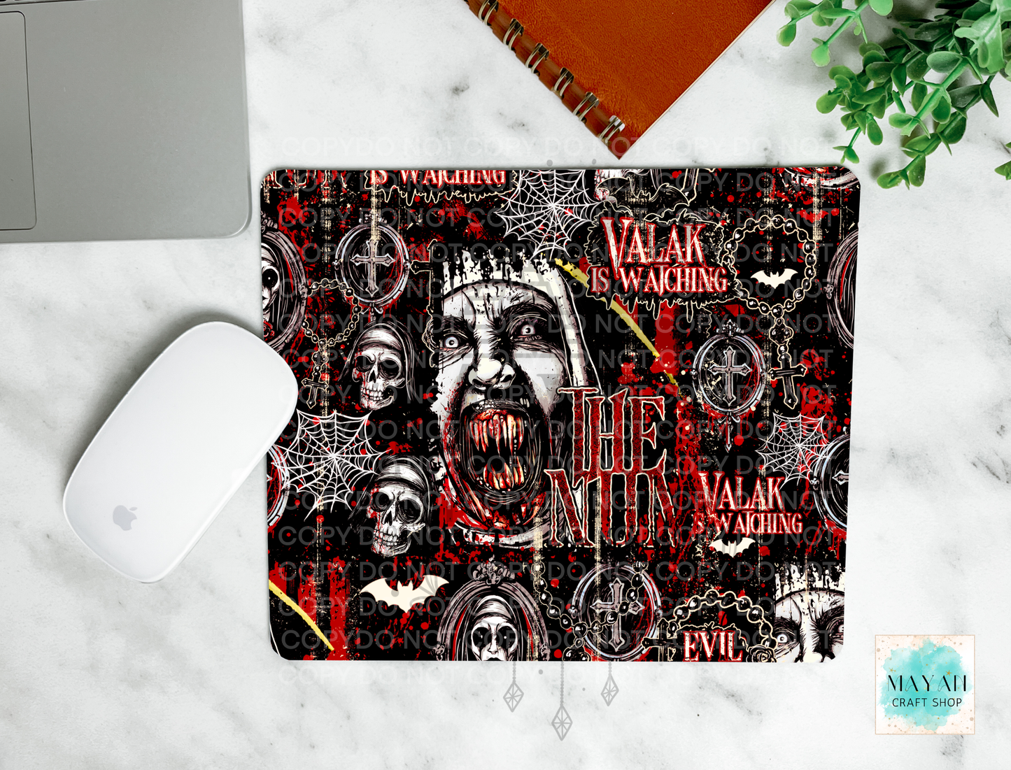 A mouse pad with a gothic horror design featuring a screaming face, skulls, bats, and a text shown on a white marble surface with a laptop mouse, notebook, and plant nearby.