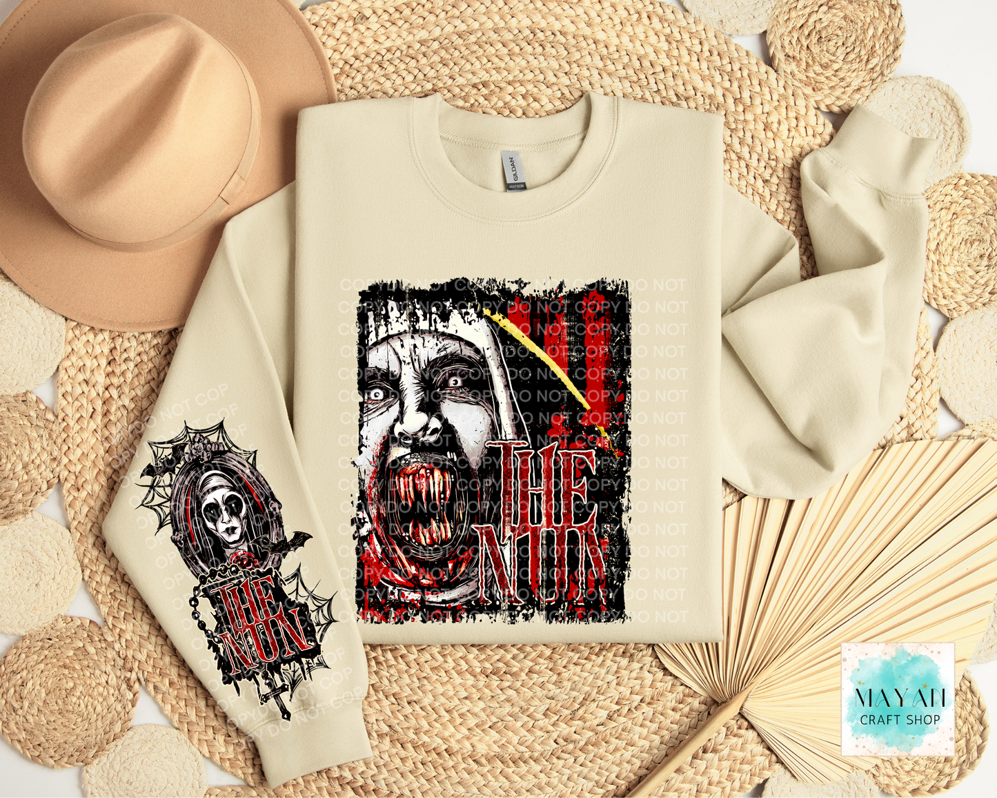A cream-colored crewneck sweatshirt with a horror-themed graphic print of a screaming face in red and black, styled in a flat-lay with a beige hat, woven placemats, and a paper fan. 
