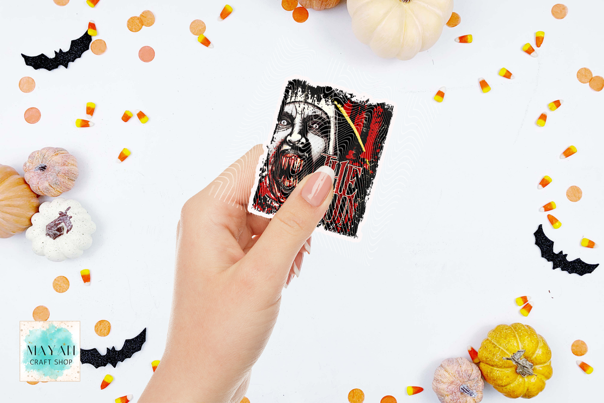 A hand holds a sticker featuring a black and red graphic illustration of a vampire. The background is a clean white surface decorated with Halloween-themed props including mini pumpkins, candy corn, and black bat cutouts.