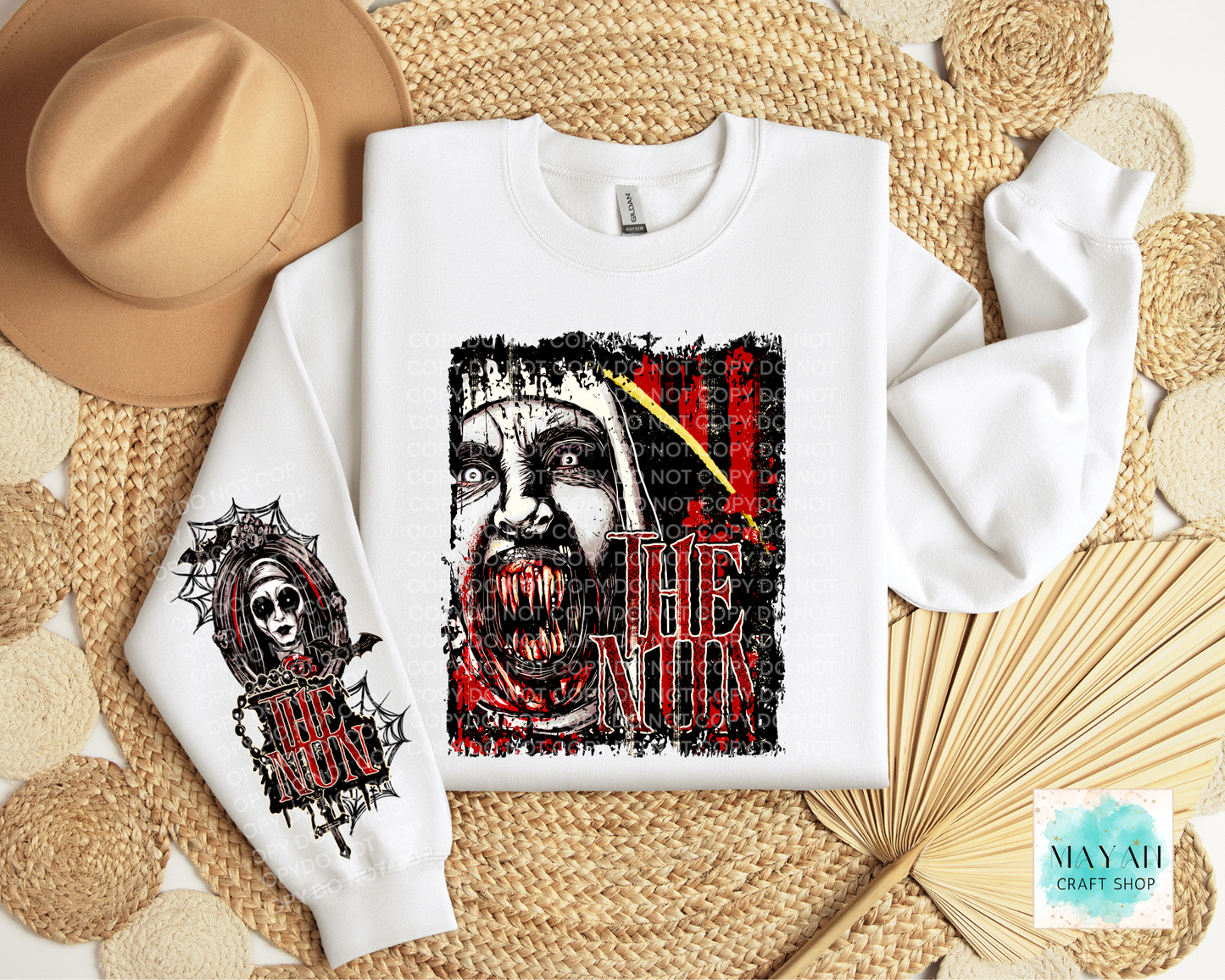 A white crewneck sweatshirt laid flat on a woven straw mat, featuring a large horror woman graphic on the front with red and black text, and a smaller matching graphic on the sleeve. A tan hat, woven mat, and dried palm leaves surround it.
