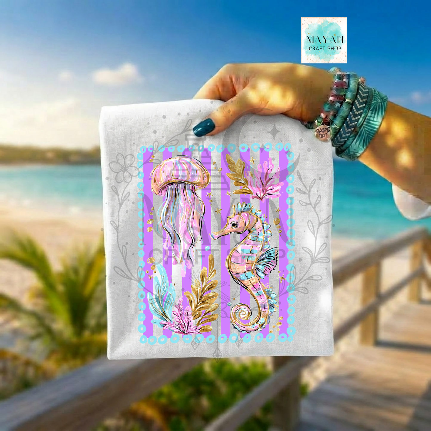 Hand holding a white shirt with pink and blue seahorse design against a beach background