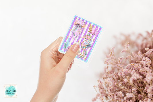 Hand holding a colorful sticker with a seahorse design against a white background with pink flowers.