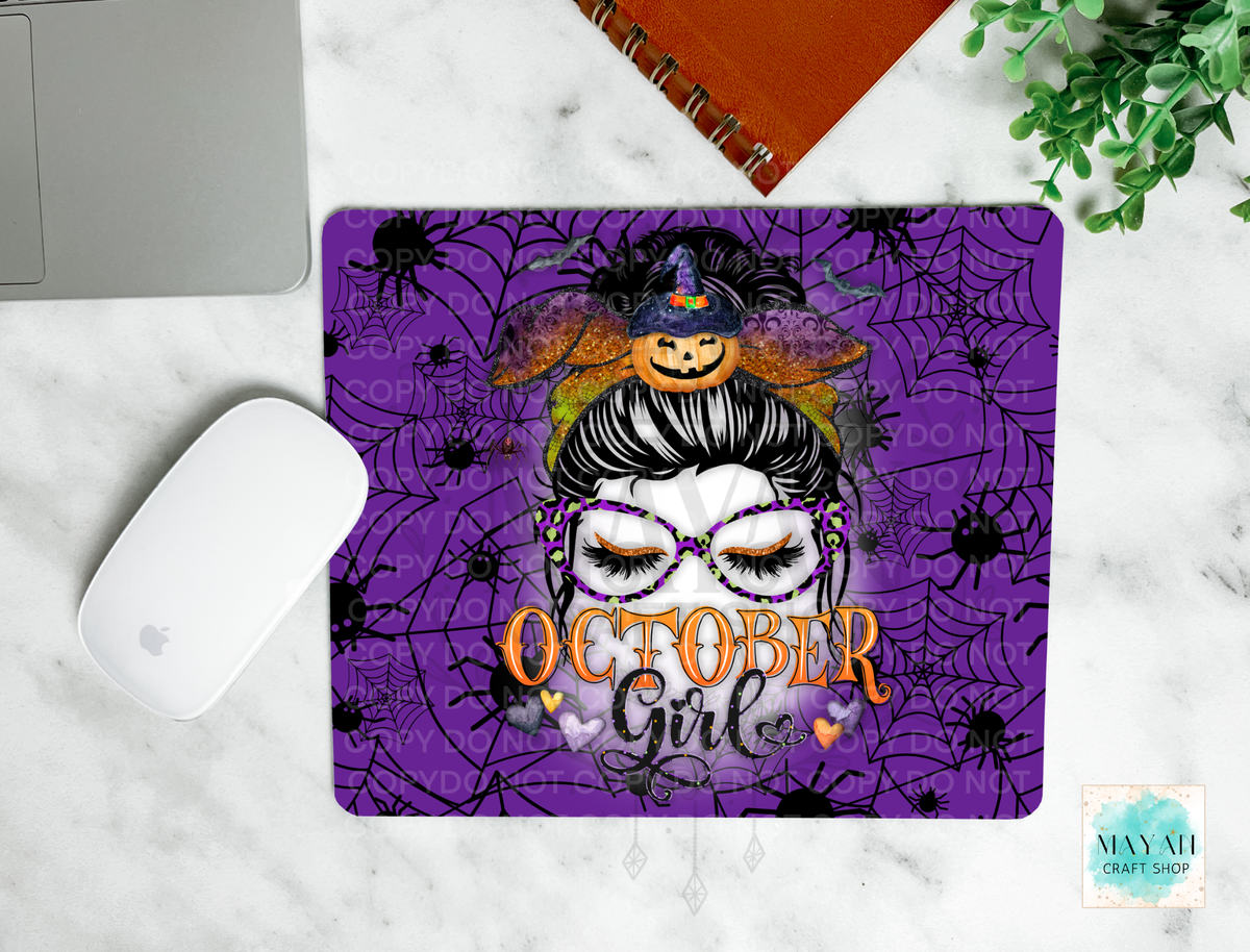 A mouse pad with a purple Halloween design featuring a woman in Mardi Gras glasses, a witch hat, spiders, and the text 'October Girls', shown on a white marble surface with a laptop, mouse, notebook, and plant nearby.