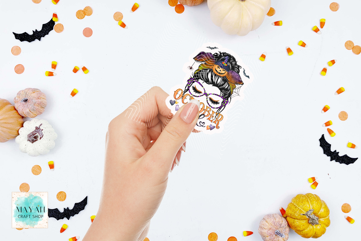 A hand holds a die-cut sticker featuring a October girl messy bun illustration of a woman with a jack-o'-lantern on her head. The background is a white flat-lay styled with small pumpkins, candy corn, and black bat cutouts.