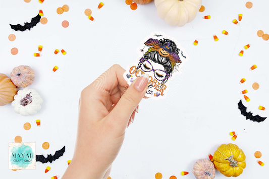 A hand holds a die-cut sticker featuring a October girl messy bun illustration of a woman with a jack-o'-lantern on her head. The background is a white flat-lay styled with small pumpkins, candy corn, and black bat cutouts.