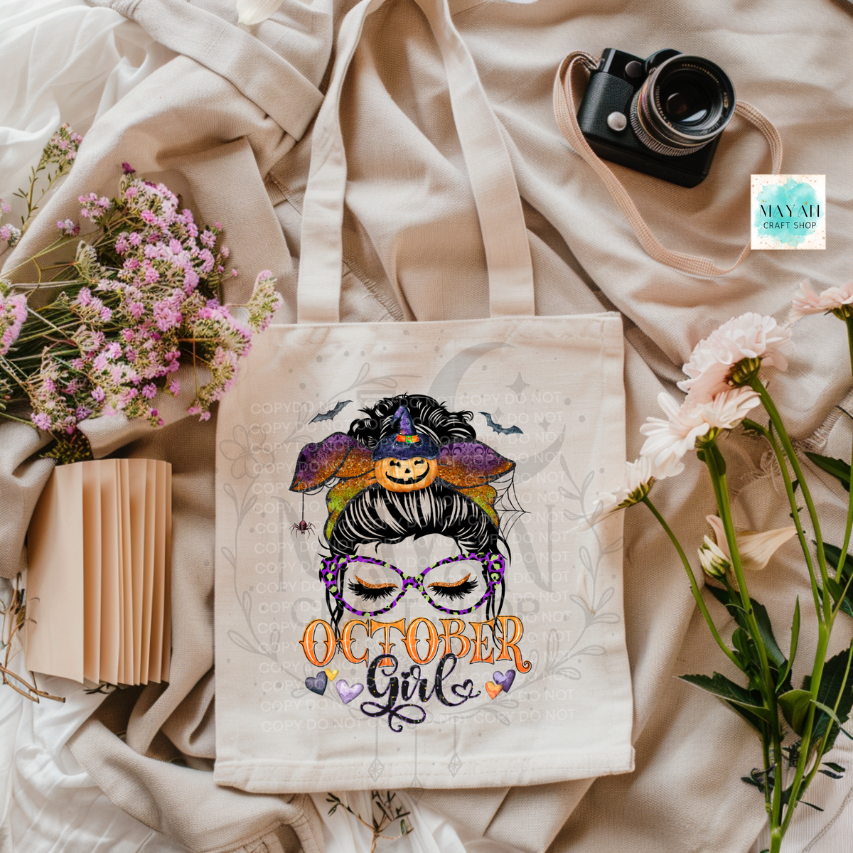A cream canvas tote bag with a Halloween-themed graphic print of a witch girl and the text 'October Girl', styled in a flat-lay arrangement with flowers, a camera, and a notebook on a beige fabric background.