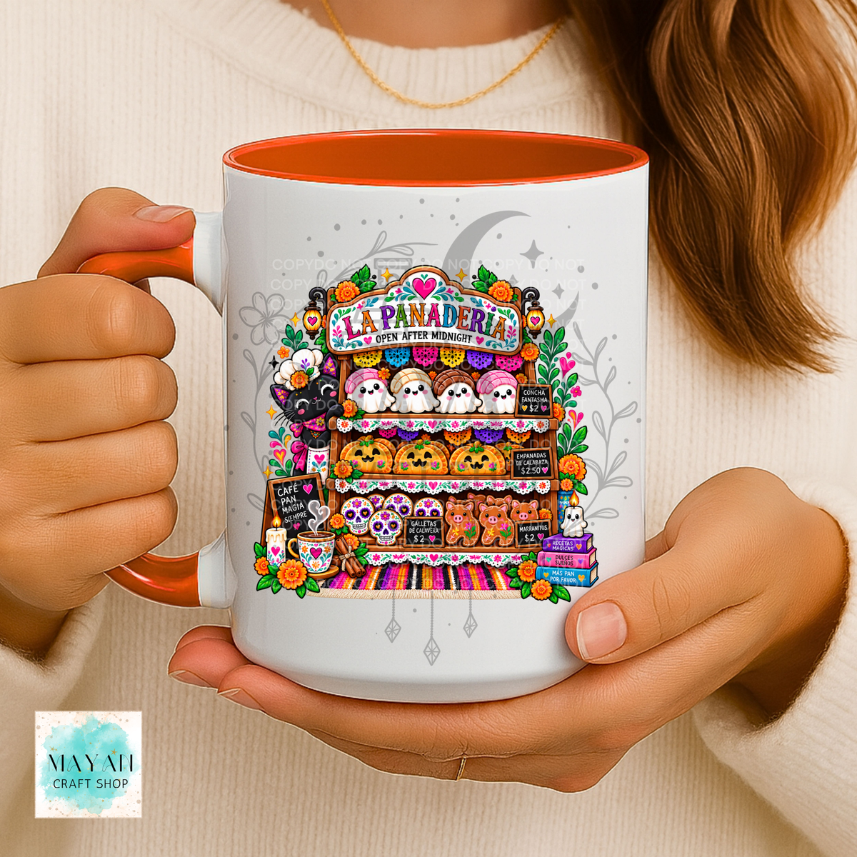 A person holds a white ceramic mug with an orange interior and handle. The mug features a colorful illustrated graphic of a Halloween-themed bakery shelf with pumpkins, ghosts, and the text 'LA PANADERIA'.