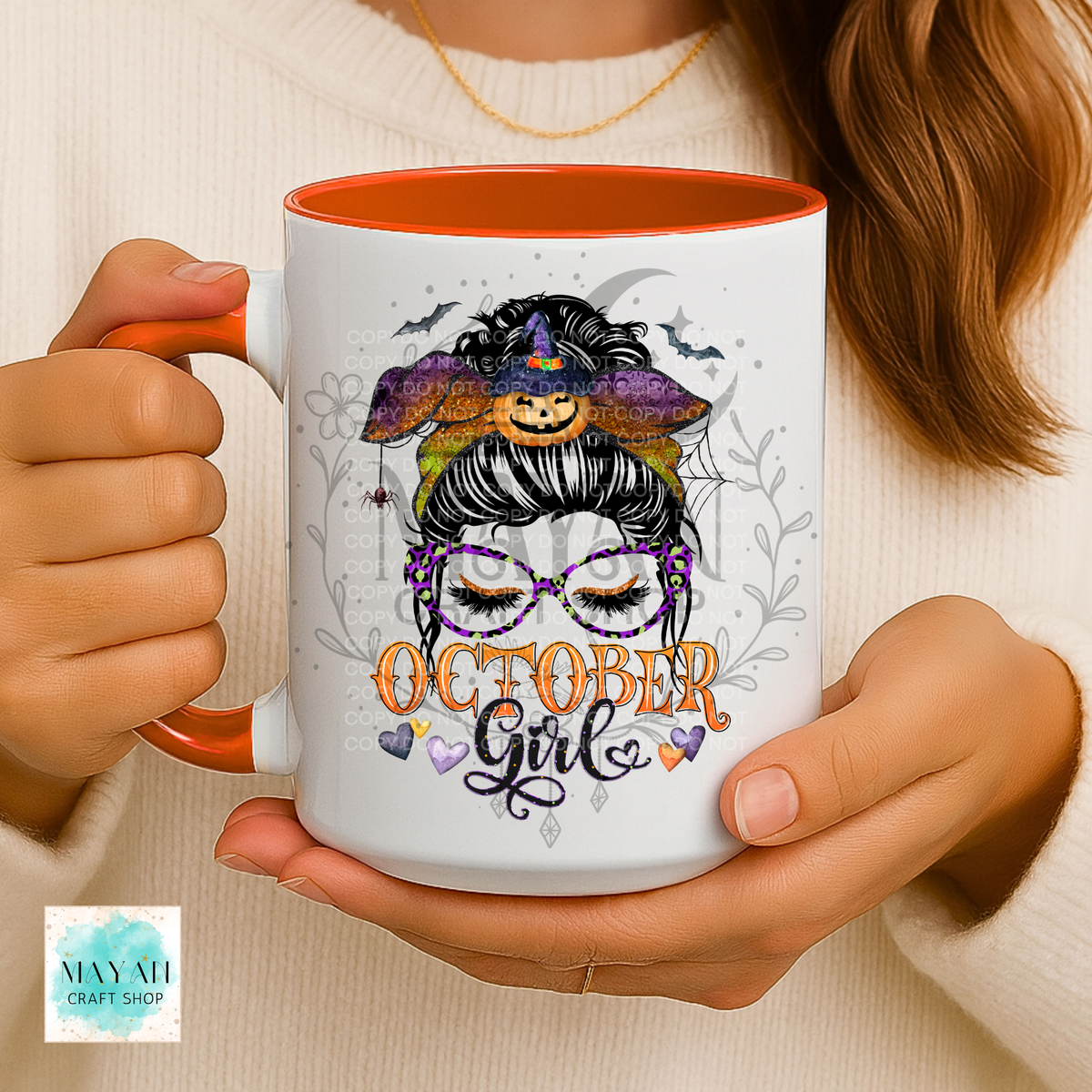 A person holds a white ceramic mug with an orange interior and handle. The mug features a graphic illustration of a woman with glasses, a witch hat with a pumpkin, and the text 'OCTOBER Girl' in orange and purple.