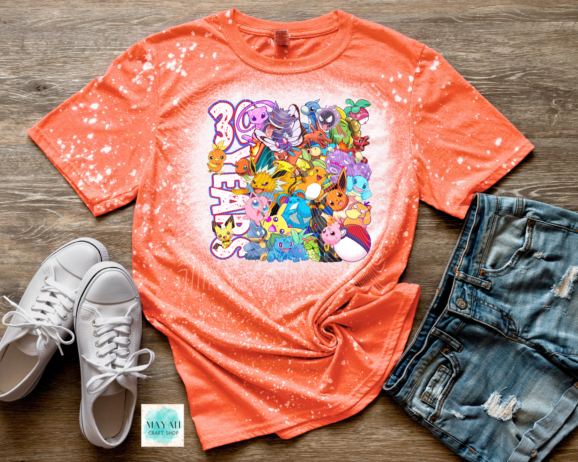 Coral t-shirt with colorful graphic design on a wooden surface with white sneakers and denim shorts.