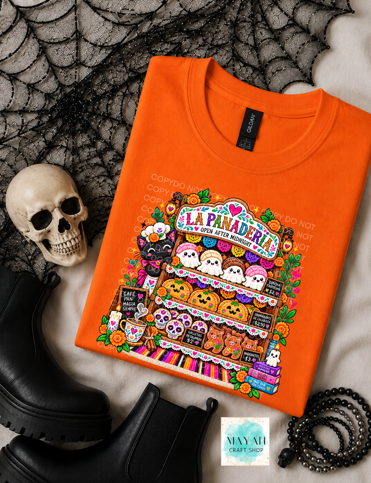An orange crewneck t-shirt with a colorful 'La Panadera' Halloween graphic, styled in a flat lay with a skull, black boots, a spiderweb, and a black beaded bracelet on a white textured background.