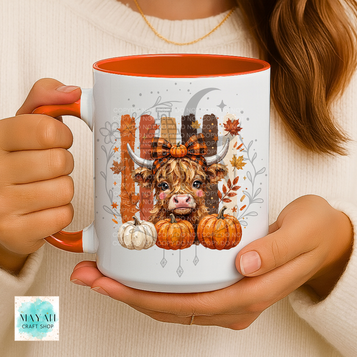 A person holds a white ceramic mug with orange interior and handle, featuring a fall-themed illustration of a shaggy Highland cow, pumpkins, leaves, and a crescent moon. The person wears a cream knit sweater.