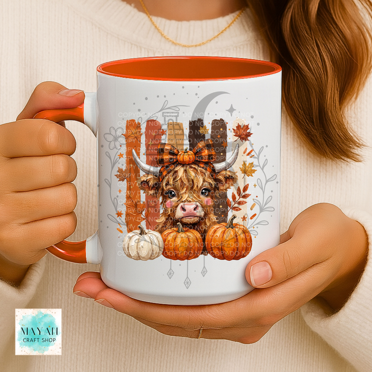 A person holds a white ceramic mug with orange interior and handle, featuring a fall-themed illustration of a shaggy Highland cow, pumpkins, leaves, and a crescent moon. The person wears a cream knit sweater.