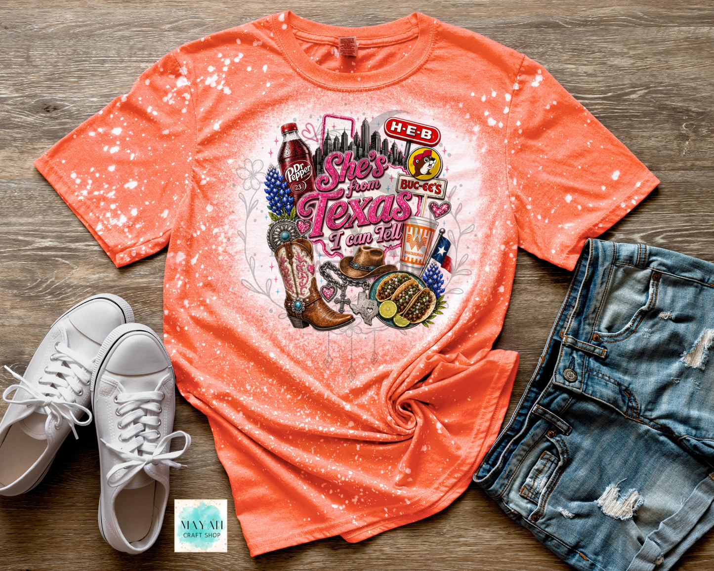 Coral t-shirt with Texas-themed graphics on a wooden surface with white sneakers and denim shorts.
