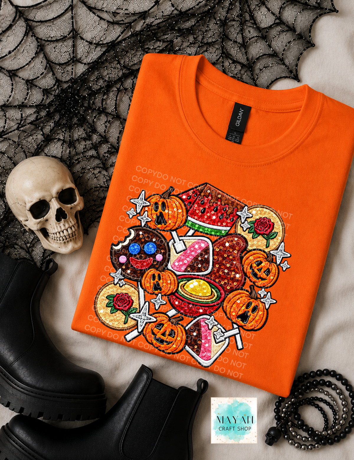 An orange crewneck shirt with a colorful Halloween-themed graphic print, styled in a flat lay with a skull, black boots, a spiderweb, and a beaded bracelet on a white textured background.
