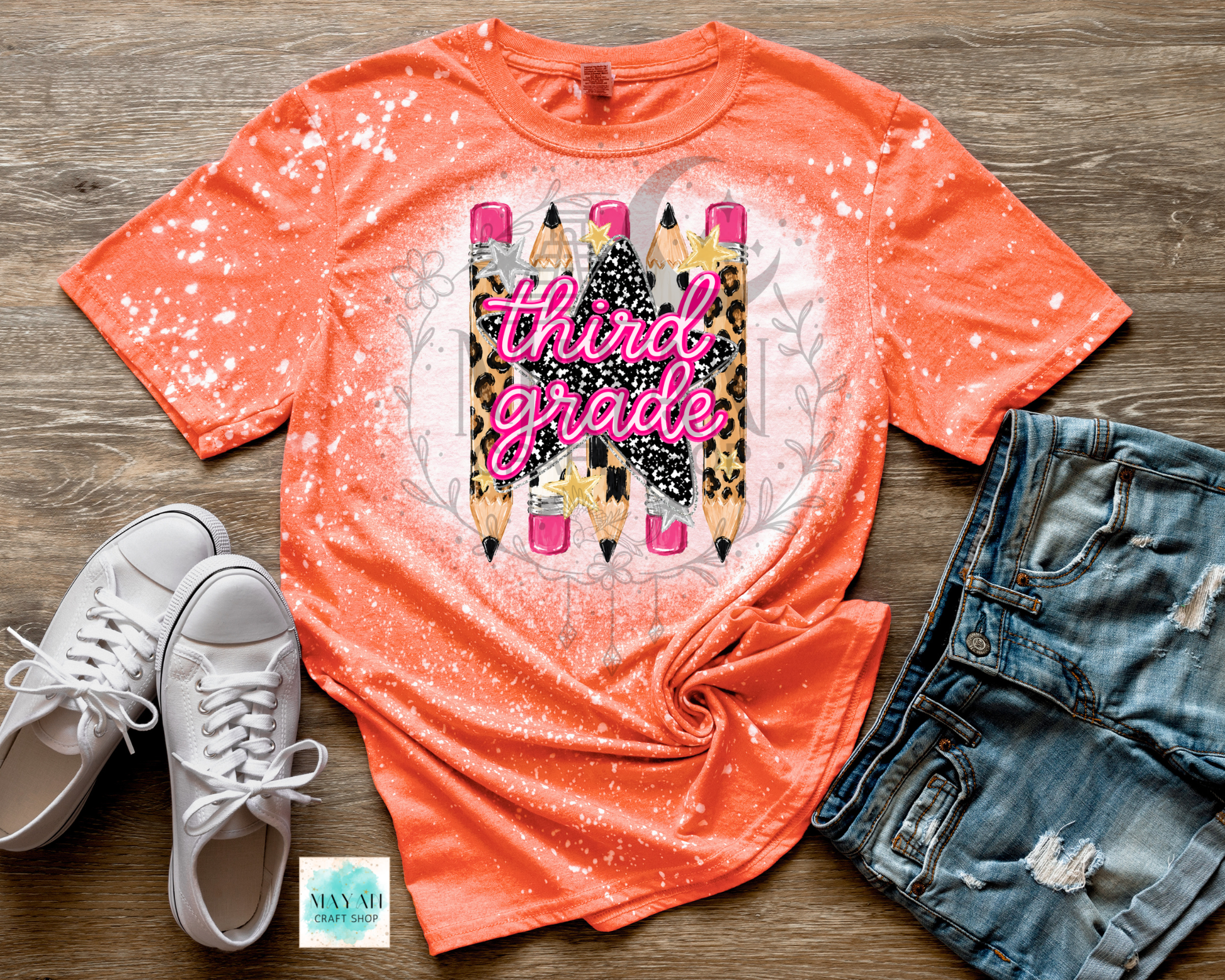 An orange short-sleeve graphic T-shirt laid flat on a wood surface, styled with white sneakers, distressed denim shorts, and a small card. The shirt has a bleached splatter effect and a central graphic of three pencils with leopard print, black glitter-style stripes, and the words 'third grade' in pink script.
