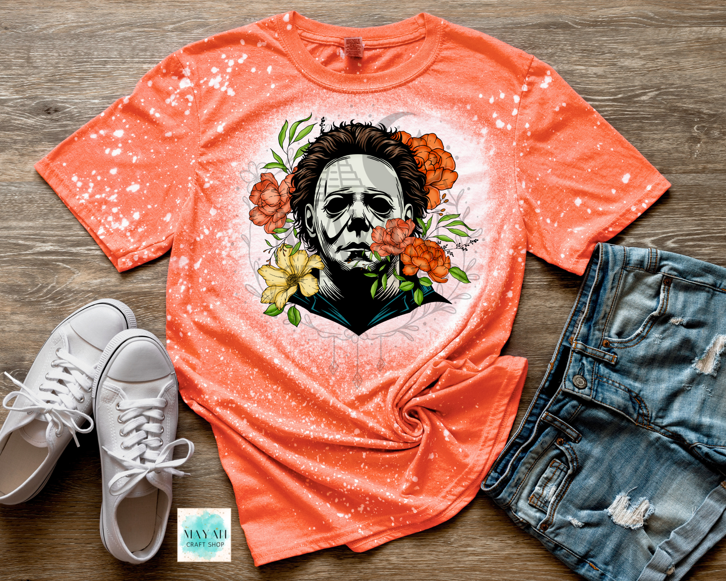 Orange t-shirt with graphic design of a face surrounded by flowers, paired with white sneakers and denim shorts on a wooden surface.