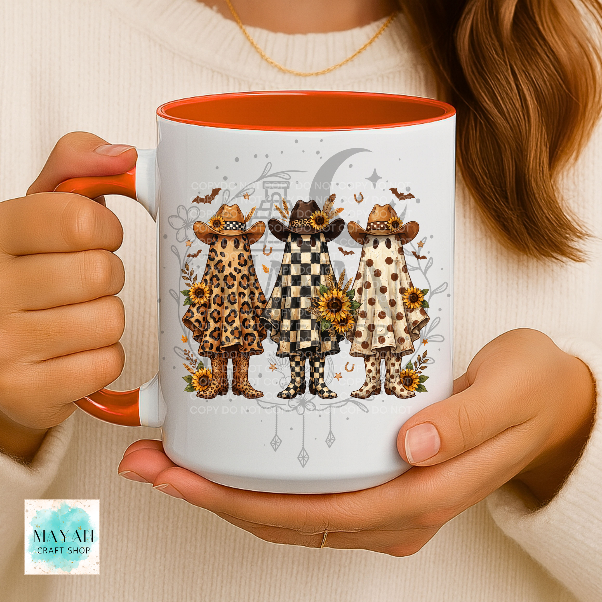 A person holds a white ceramic mug with a bright orange interior and handle. The mug features a printed illustration of three stylized figures in cowboy hats and boots, each in a different pattern (leopard print, black-and-white check, beige polka dots), with a gray crescent moon, sunflowers, and bats in the background.