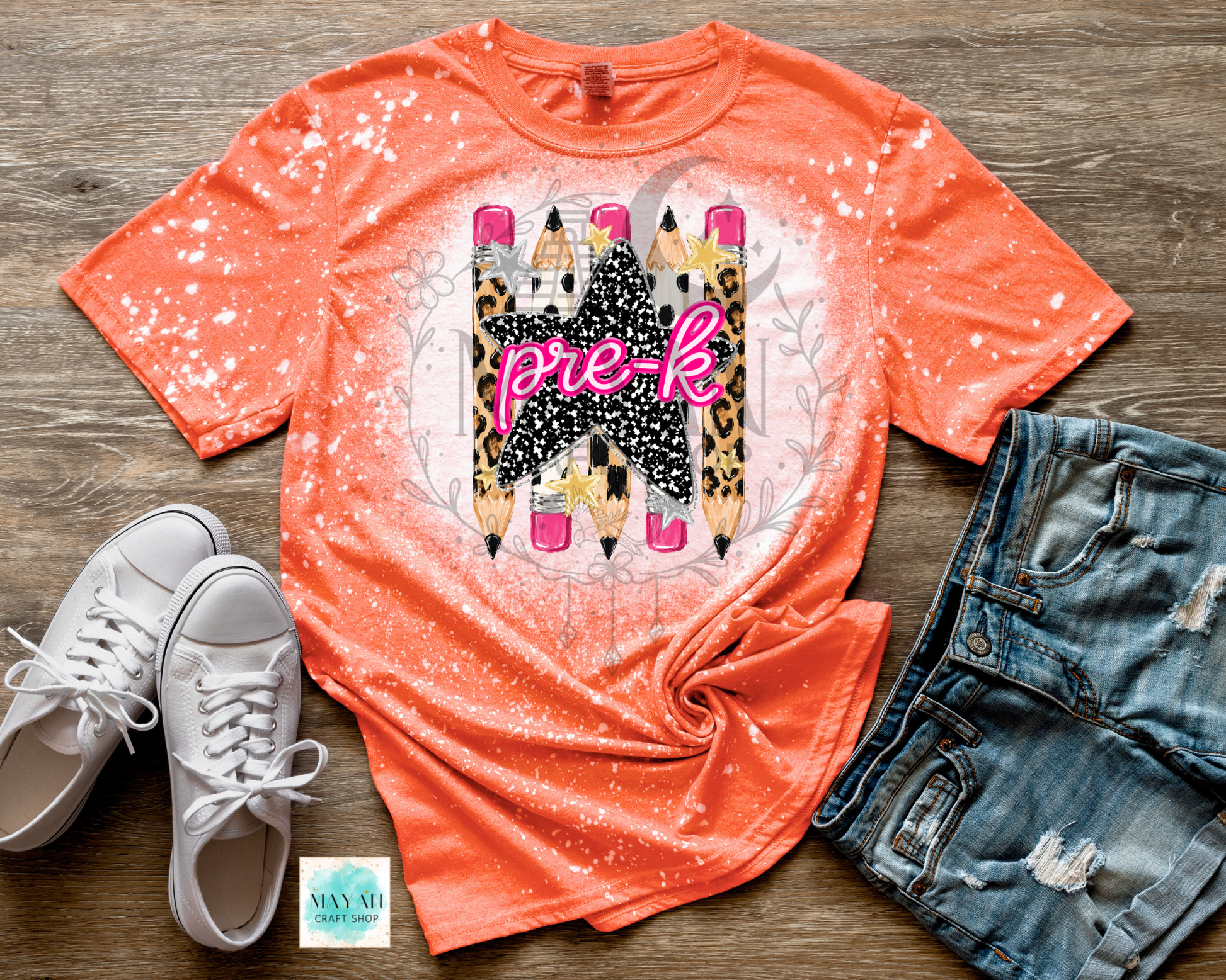 An orange short-sleeve graphic T-shirt laid flat on a wood surface, styled with white sneakers, distressed denim shorts, and a small card. The shirt has a white bleach-splatter effect and a central graphic of three pencils with leopard print, black glitter, and pink 'pre-k' lettering.