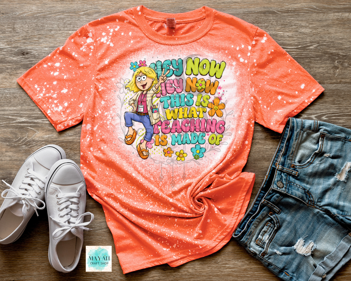 An orange short-sleeve graphic T-shirt laid flat on a wood surface, styled with white sneakers, distressed denim shorts, and a small card. The shirt has a white bleach-splatter effect and a colorful cartoon-style graphic with the text 'TEACH NOW TEACH NOW THIS IS WHAT TEACHING IS MADE OF.